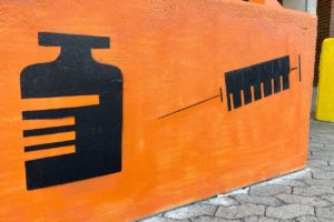 Impfzentrum Hürth Darstellung Spritze und Viole auf Betonblock vor Gebäude